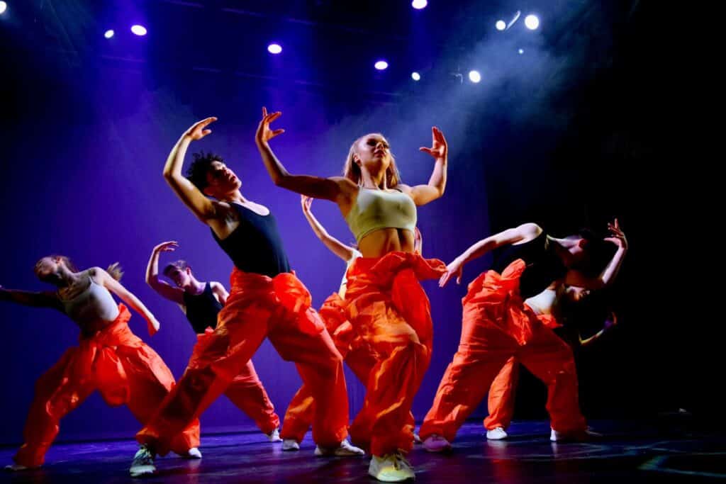 The college students from Redbridge Drama and Dance course perform a dance on stage in orange trousers and cream and black tops