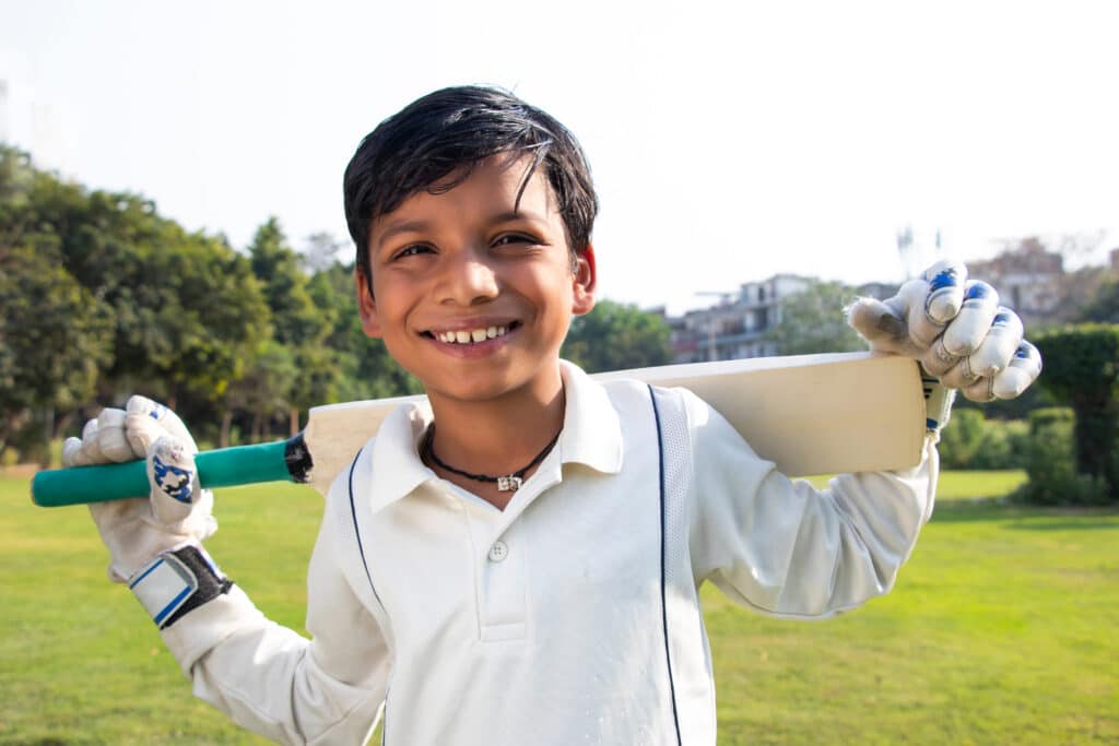 Boy in a park wearing cricket gear, holding a cricket bat behind his back and smiling at the camera